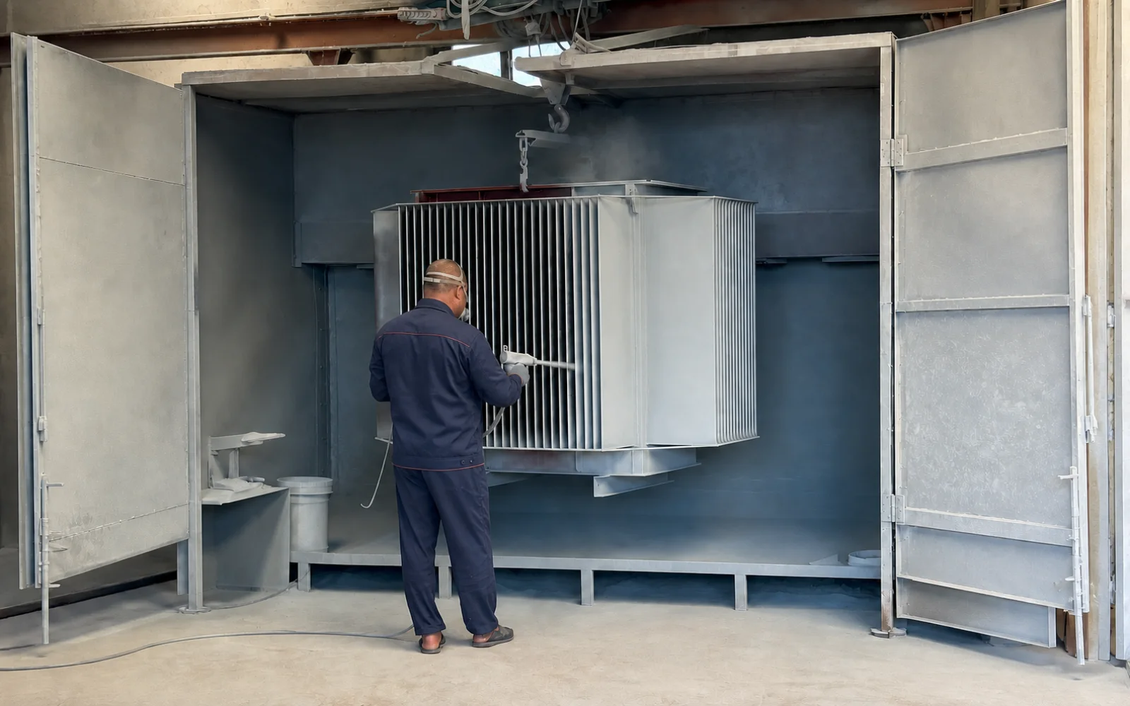 Worker in factory uniform spray coating a transformer tank in the booth